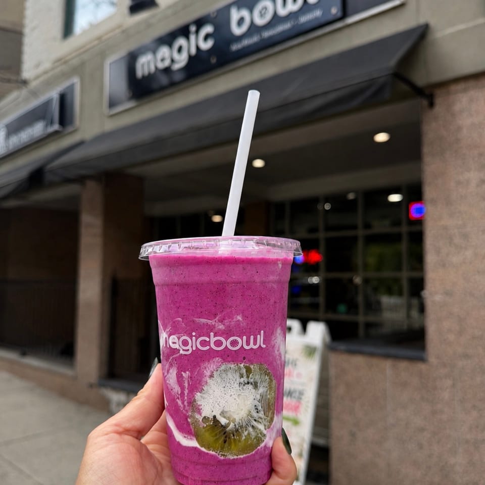 Hand holds a purple kiwi smoothie outside Magic Bowl, with the restaurant storefront and sign visible behind it.
