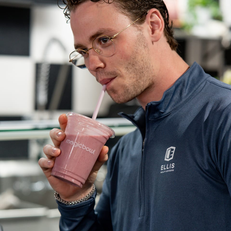 Man wearing glasses drinks a pink smoothie through a straw inside a bright café.