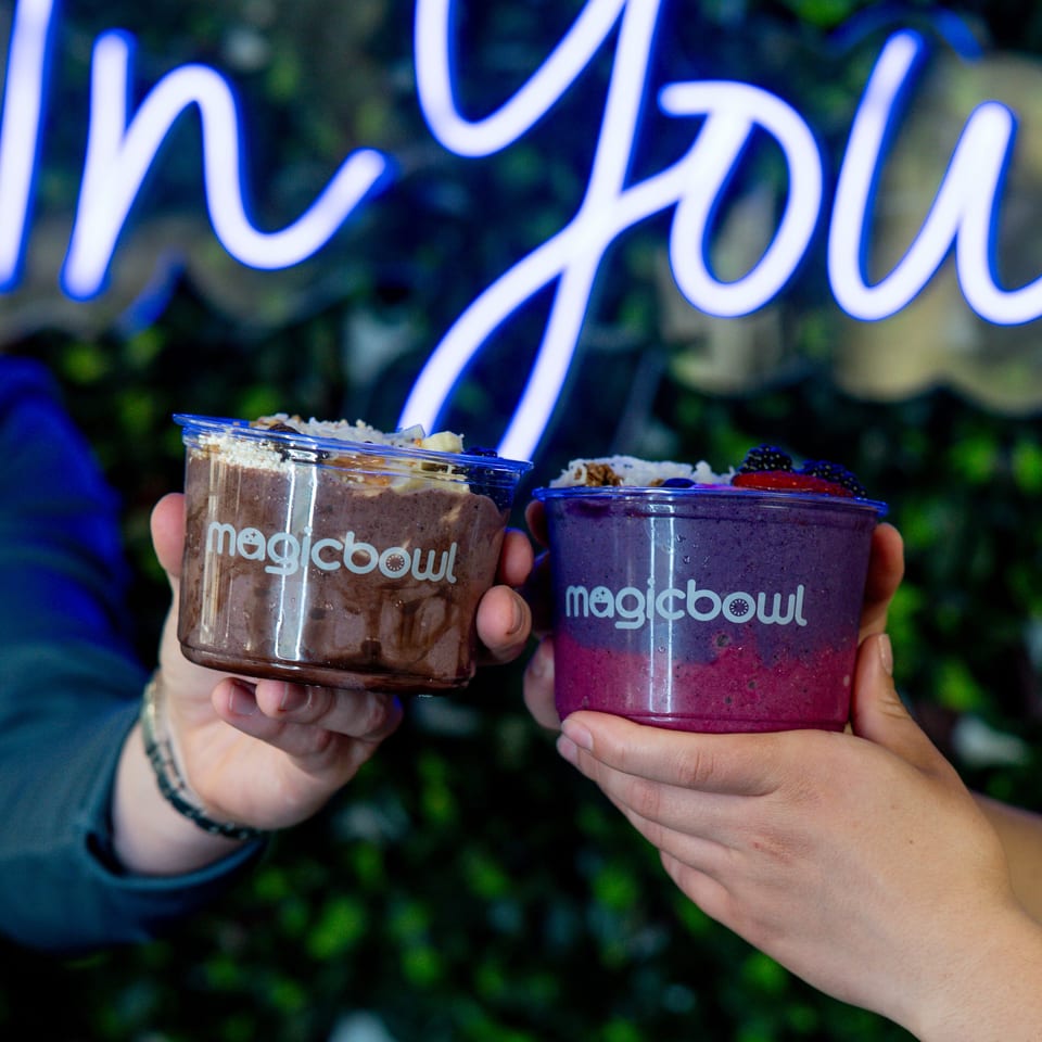 Two hands hold topped chocolate and berry smoothie bowls in Magicbowl cups beneath a glowing neon sign.