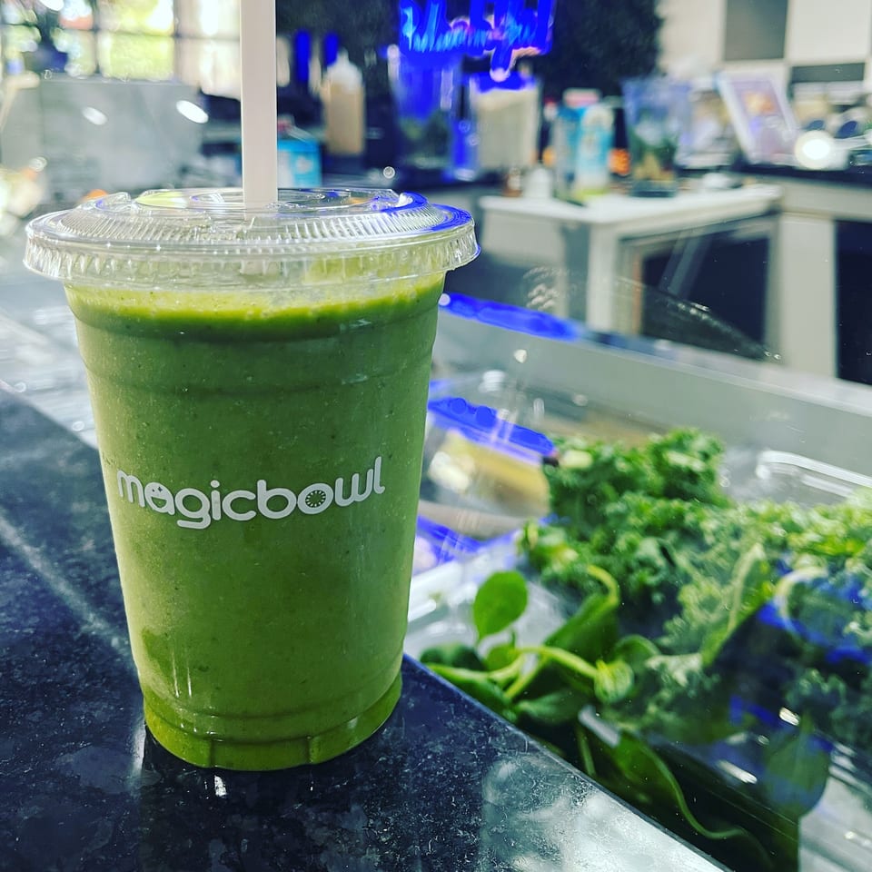 A Magic Bowl green smoothie with a straw, next to fresh greens in a display case.
