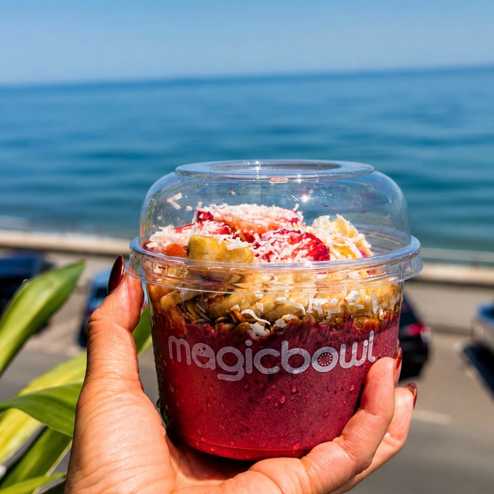 Hand holding a layered Magic Bowl smoothie topped with fruit and coconut, with the ocean visible in the background.
