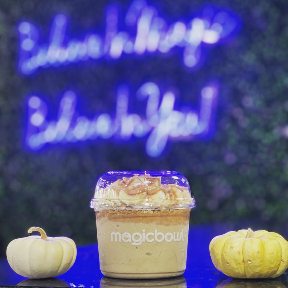Magic Bowl smoothie with banana and peanut butter, flanked by two small pumpkins, under a blue neon sign.