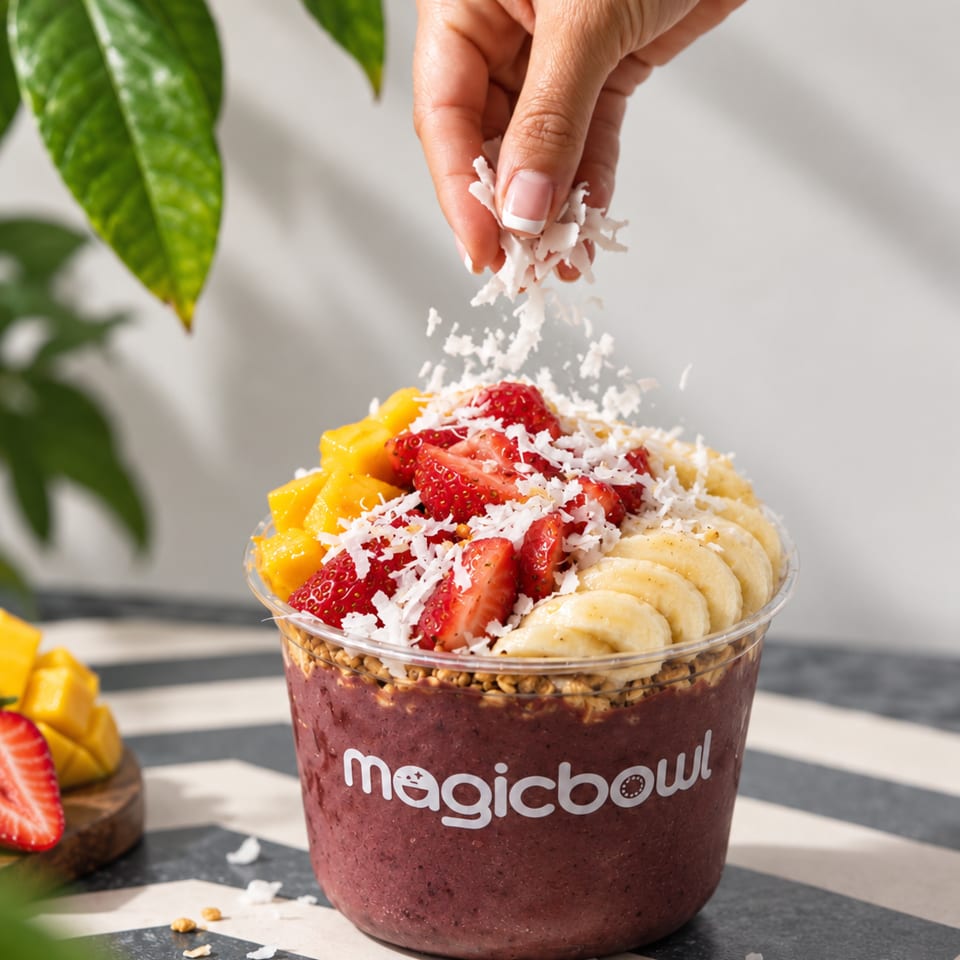 Magicbowl acai bowl topped with strawberries, banana, mango, granola, and shredded coconut as a hand sprinkles coconut.