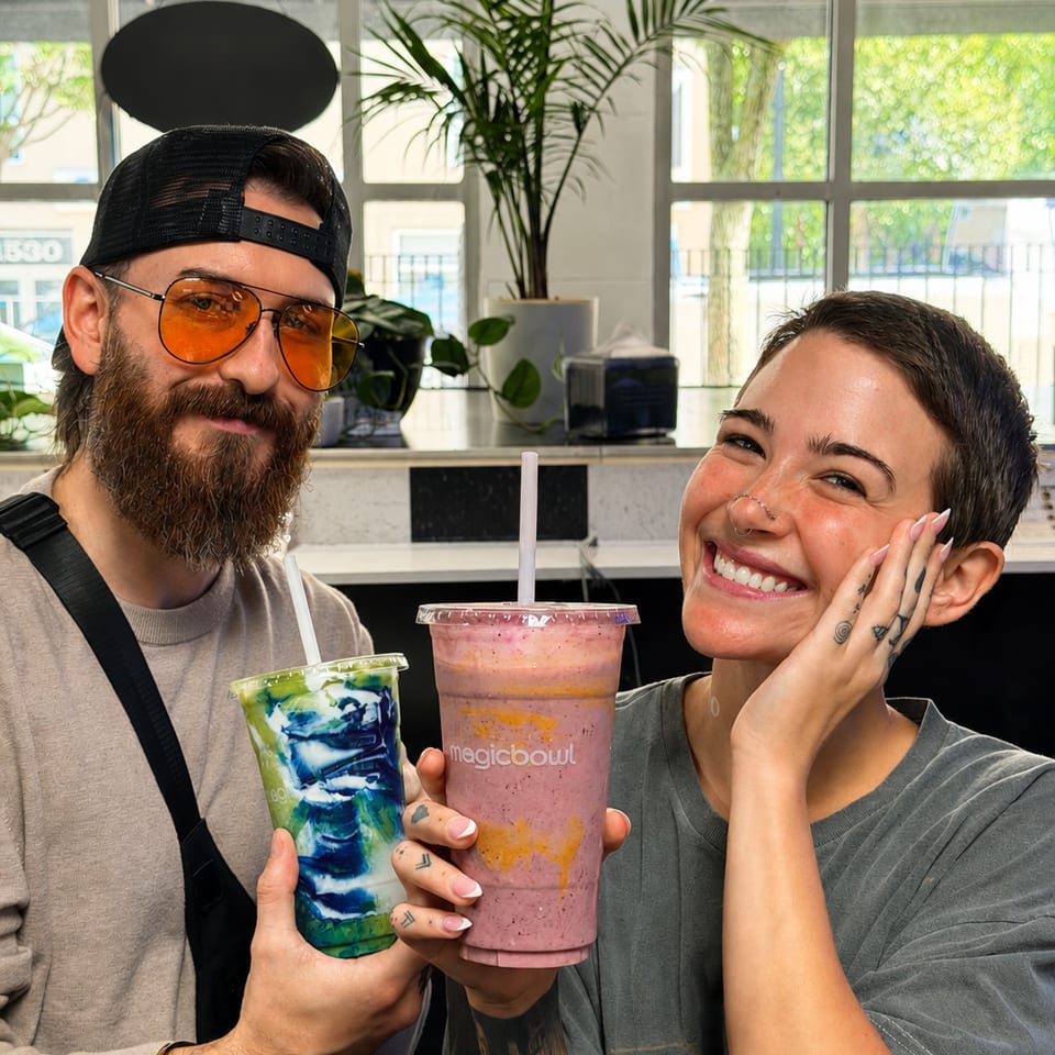 Two smiling customers hold colorful smoothies inside a bright café with plants and large windows.