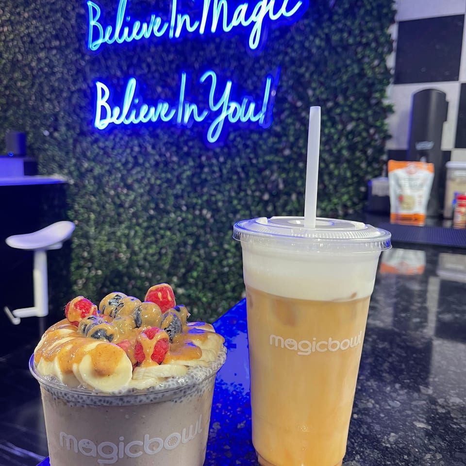 Magic Bowl smoothie bowl with fruit and a drink with cream top, under a neon sign.